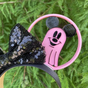 May include: Pink Mickey Mouse ghost headband with black sequin bow. The headband is made of plastic and features a pink ghost with Mickey Mouse ears and a black outline. The bow is made of black sequins and is attached to the headband.