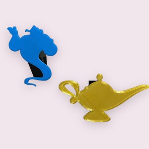 May include: A blue genie silhouette and a gold lamp silhouette, both with black bases. These are 3D printed ornaments.