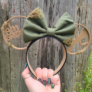May include: A headband featuring a large olive green fabric bow adorned with patches of green moss. The circular ears are made of tan plastic, with the left ear displaying the text I AM GROOT and the right ear featuring a silhouette of a small tree-like character.