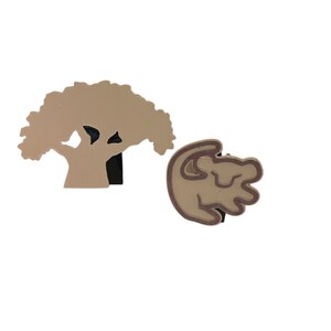 May include: A set of two brown and tan felt ornaments. One ornament is a silhouette of a tree with a rounded canopy. The other ornament is a silhouette of a lion cub curled up.