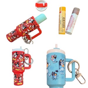 K&ouml;nnte beinhalten: Eine Sammlung von neuartigen Lippenbalsam-Beh&auml;ltern und -Tuben. Ein roter Beh&auml;lter zeigt ein Mickey Mouse-Muster, w&auml;hrend ein anderer blau mit Bluey-Figuren ist. Au&szlig;erdem sind zwei Lippenbalsam-Tuben abgebildet.