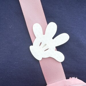 Puede incluir: Un bolso de hombro rosa y blanco con un dije de guante de Mickey Mouse blanco. El bolso tiene cierre de cremallera.