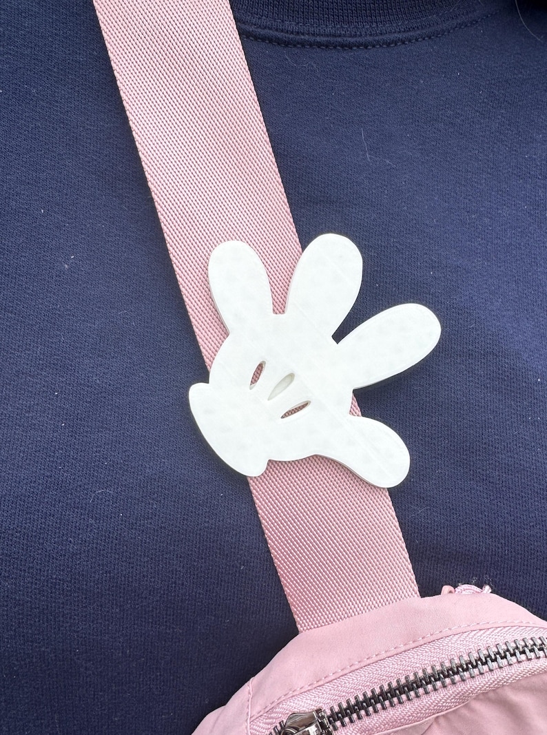 Puede incluir: Un bolso de hombro rosa y blanco con un dije de guante de Mickey Mouse blanco. El bolso tiene cierre de cremallera.