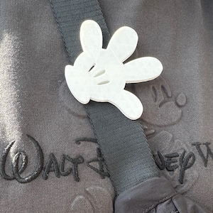Puede incluir: Un amuleto de guante blanco de Mickey Mouse sujeto a una correa de hombro negra. La correa est&aacute; unida a una bolsa negra. La bolsa est&aacute; sobre una sudadera gris con el texto "Walt Disney World" bordado.