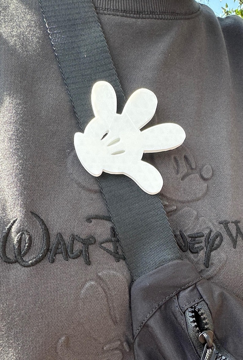 Puede incluir: Un amuleto de guante blanco de Mickey Mouse sujeto a una correa de hombro negra. La correa est&aacute; unida a una bolsa negra. La bolsa est&aacute; sobre una sudadera gris con el texto "Walt Disney World" bordado.