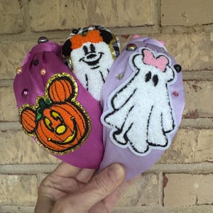 May include: Three Halloween-themed headbands. One is purple with a jack-o'-lantern, one is white with a cartoon ghost, and the other is white with a cartoon character. Each headband has decorative embellishments.