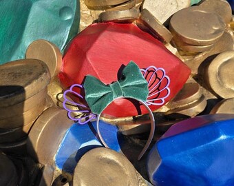 May include: A green velvet bow headband with a purple seashell design. The headband is set against a backdrop of gold coins, red and blue gems, and green gems.