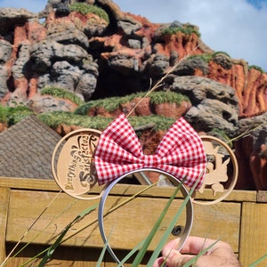 May include: A pair of white mouse ears with a red and white gingham bow. The ears are decorated with a wooden cutout that says "It's a Small World" and features a whimsical design.