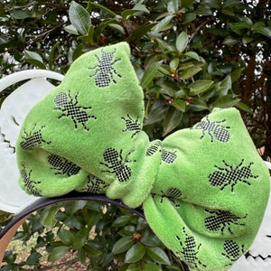 Pode incluir: Orelhas de Minnie Mouse de veludo verde com um padr&atilde;o de formigas pretas. O la&ccedil;o &eacute; grande e felpudo.