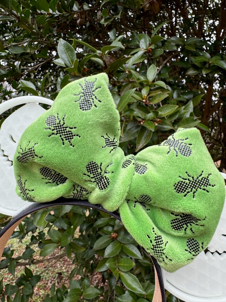 Pode incluir: Orelhas de Minnie Mouse de veludo verde com um padr&atilde;o de formigas pretas. O la&ccedil;o &eacute; grande e felpudo.