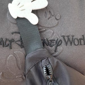 Puede incluir: Una ri&ntilde;onera negra con dos cremalleras y un recorte blanco en forma de mano de Mickey Mouse sujeto a la correa. La ri&ntilde;onera se lleva sobre una camiseta gris con el texto "Walt Disney World" bordado.