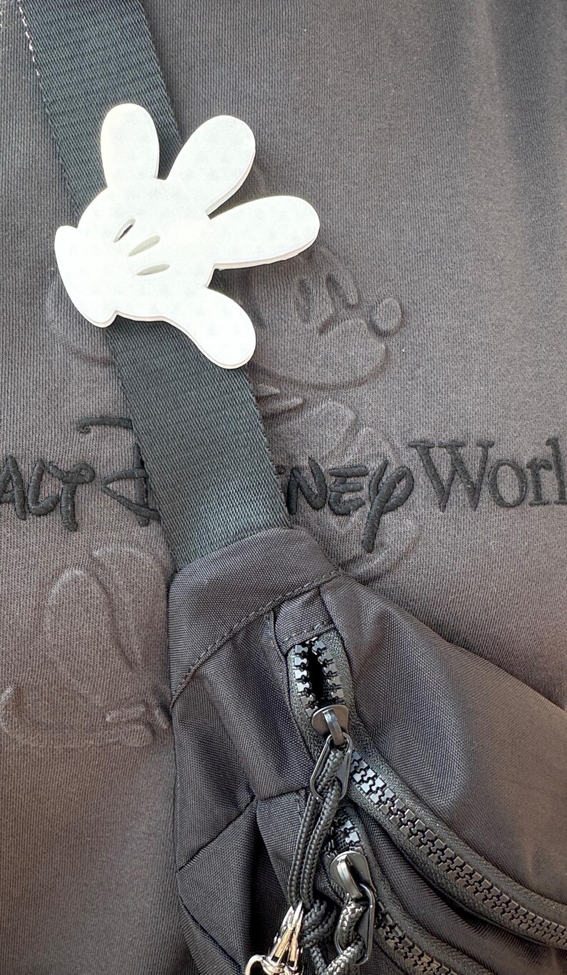Puede incluir: Una ri&ntilde;onera negra con dos cremalleras y un recorte blanco en forma de mano de Mickey Mouse sujeto a la correa. La ri&ntilde;onera se lleva sobre una camiseta gris con el texto "Walt Disney World" bordado.