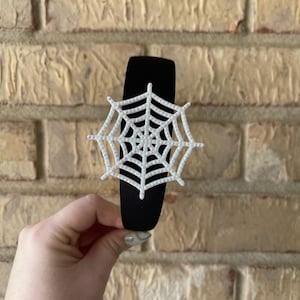 May include: Black headband with a white spiderweb design. The spiderweb is centered on the headband. The headband is made of a soft material. The background is a brick wall.