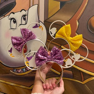 May include: A pair of white Minnie Mouse ears with a silhouette of Mrs. Potts and Chip from Beauty and the Beast. The ears are decorated with purple and yellow sequined bows.