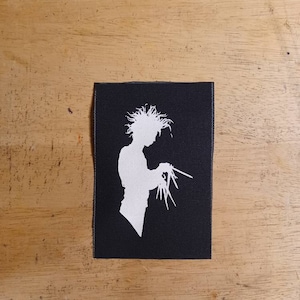 Può includere: Silhouette bianca di una persona con molti capelli che tiene un paio di forbici su un pezzo di stoffa nero.