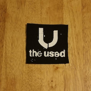 Puede incluir: Parche cuadrado negro con el logotipo blanco de la banda "U" sobre "the used" en una fuente desgastada. El parche está sobre una superficie de madera.