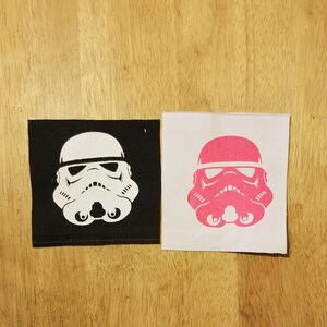 May include: Two square fabric patches featuring a Stormtrooper helmet design. One patch is black with a white helmet, and the other is white with a pink helmet. The patches are placed on a wooden surface.