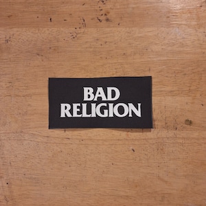 Puede incluir: Un parche rectangular negro con las palabras "BAD RELIGION" en letras blancas y en negrita. El parche está centrado sobre una superficie de madera.