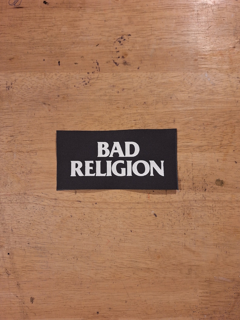 May include: A black rectangular patch with the words "BAD RELIGION" in bold white capital letters. The patch is centered on a wooden surface.