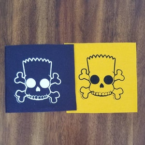 May include: Two iron-on patches featuring a skull with a cartoon character's hair. One patch is on a dark blue background and the skull is white. The other patch is on a yellow background and the skull is black.