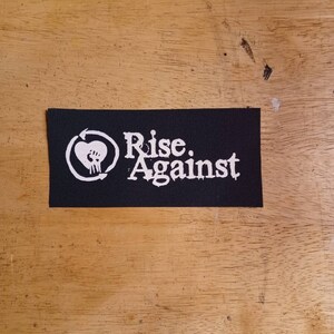 May include: A black rectangular patch featuring the words "Rise. Against" in white, accompanied by a white heart and fist graphic. The patch is displayed on a wooden surface.