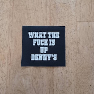 May include: Black square patch with white text that reads "WHAT THE FUCK IS UP DENNY'S". The patch is on a light brown wooden surface. The text is in a bold, sans-serif font.