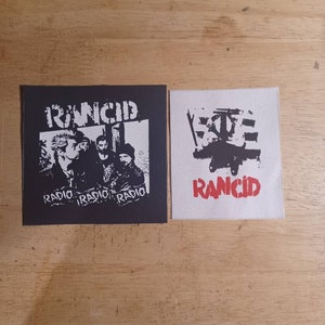 Puede incluir: Dos parches cuadrados con el nombre de la banda "Rancid". Uno es negro con una foto de la banda y la palabra "Radio" repetida. El otro es blanco con un gráfico y el nombre de la banda en rojo.