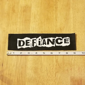May include: Black and white woven patch with the word "DEFIANCE" in white letters with a distressed, torn look. The patch is approximately 3.5 inches wide and 1 inch tall.