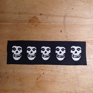 May include: Black rectangular patch featuring five white skull graphics. The skulls have a distinctive design with large eye sockets and a wide grin. The patch is likely for clothing or accessories.