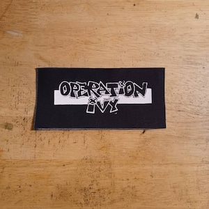 Pode incluir: Um patch retangular preto com as palavras "OPERATION IVY" em branco, com uma fonte de estilo grafite. O texto está centrado em uma barra horizontal branca. O patch está em uma superfície de madeira.
