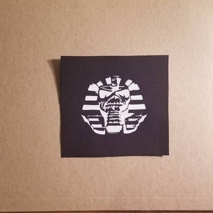May include: A black square patch with a white graphic of a pharaoh's head. The design includes a stylized face and headdress. The patch is centered on a black background. It is likely intended for application on clothing or accessories.