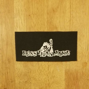 以下が含まれることがあります： 白文字で「LESS THAN JAKE」と漫画のイラストが描かれた黒い長方形のパッチ。イラストは、とがった髪の毛のキャラクターと棒人間の姿です。パッチは木製の表面にあります。