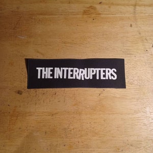 May include: A black rectangular patch with the white text "THE INTERRUPTERS". The patch is likely made of fabric and is displayed on a wooden surface. The text is in a bold, sans-serif font.