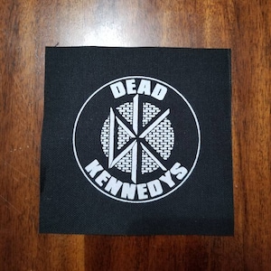 Może przedstawiać: Czarna łatka z tkaniny z białym okręgiem zawierającym tekst "DEAD KENNEDYS" i stylizowanym białym symbolem w środku.
