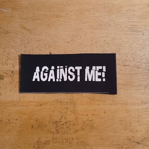 May include: A black rectangular patch with the words "AGAINST ME!" in white, stylized text. The patch is made of a woven material and is set against a wooden background.