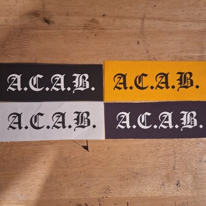Op de afbeelding: Vier stoffen patches met de tekst "A.C.A.B." in zwarte gotische letters. De patches zijn in zwart, wit, grijs en geel.