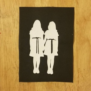 May include: A black patch featuring a white silhouette of two figures holding hands. The figures are wearing dresses and have long hair. The patch is rectangular and has a stitched border.