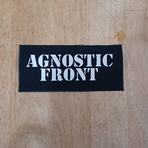 Puede incluir: Un parche rectangular negro con el texto blanco "AGNOSTIC FRONT". El parche es probablemente de tela y se muestra sobre una superficie de madera de color marrón claro. El texto está en negrita, fuente sans-serif.