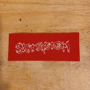 Könnte beinhalten: Ein rechteckiger roter Aufnäher mit dem weißen Text "SKIT SYSTEM" in einer stilisierten, tropfenden Schriftart. Der Aufnäher besteht aus gewebtem Material und wird auf einer Holzoberfläche präsentiert.