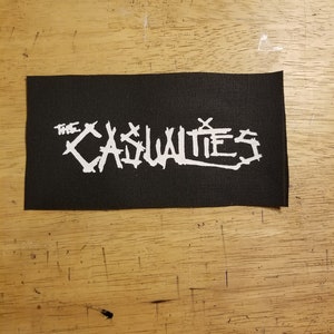 Puede incluir: Parche rectangular negro con el texto blanco "THE CASUALTIES" en una fuente estilizada. El parche es probablemente de tela y está diseñado para coser o planchar sobre ropa o accesorios. El parche está sobre una superficie de madera.