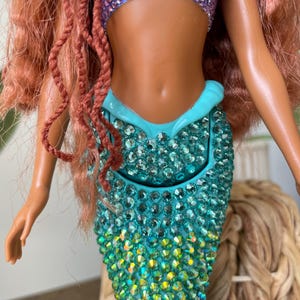 May include: A mermaid doll with a sparkling, jewel-encrusted teal tail that transitions to purple. The doll has a purple sequined top and red hair. The doll's skin is a tan color. The doll is a toy.