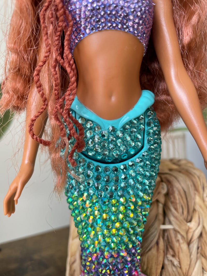 May include: A mermaid doll with a sparkling, jewel-encrusted teal tail that transitions to purple. The doll has a purple sequined top and red hair. The doll's skin is a tan color. The doll is a toy.