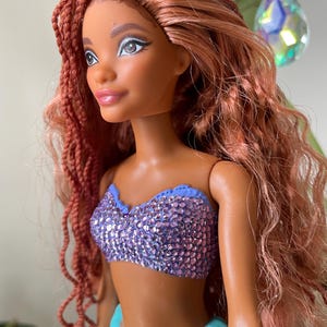 May include: A doll with auburn hair styled in braids and loose waves. The doll has brown skin, and is wearing a sparkly purple top and a turquoise skirt. The doll's eye makeup is silver. The doll is a mermaid.