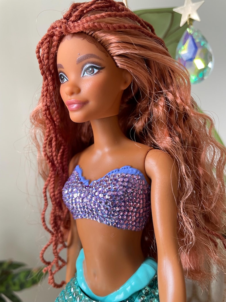 May include: A doll with auburn hair styled in braids and loose waves. The doll has brown skin, and is wearing a sparkly purple top and a turquoise skirt. The doll's eye makeup is silver. The doll is a mermaid.