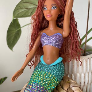 May include: A mermaid doll with brown skin, red braided hair, and a purple sequined top. The doll's tail is a gradient of green, blue, and pink sequins. The doll is posed with one arm raised, and a white star is attached to the doll's hair.