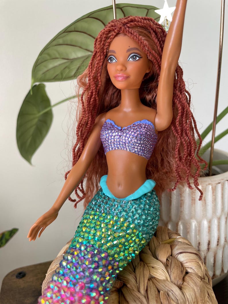 May include: A mermaid doll with brown skin, red braided hair, and a purple sequined top. The doll's tail is a gradient of green, blue, and pink sequins. The doll is posed with one arm raised, and a white star is attached to the doll's hair.