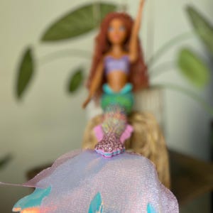 May include: A mermaid doll with red hair, a purple top, and a green tail. The doll is wearing a sparkly pink and blue accessory. The doll is set against a blurred background with green leaves.