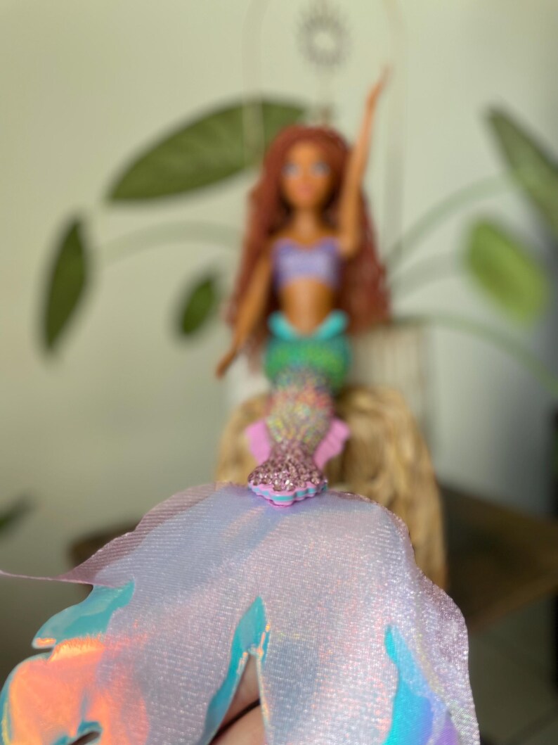 May include: A mermaid doll with red hair, a purple top, and a green tail. The doll is wearing a sparkly pink and blue accessory. The doll is set against a blurred background with green leaves.