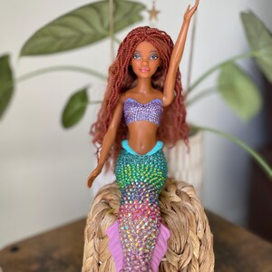 May include: A mermaid doll with long auburn hair and a sparkling, multi-colored tail. The doll wears a purple sequined top and has one arm raised. The doll is seated on a woven basket, with a plant in the background.