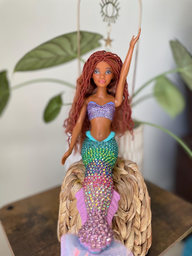 May include: A mermaid doll with long auburn hair and a sparkling, multi-colored tail. The doll wears a purple sequined top and has one arm raised. The doll is seated on a woven basket, with a plant in the background.